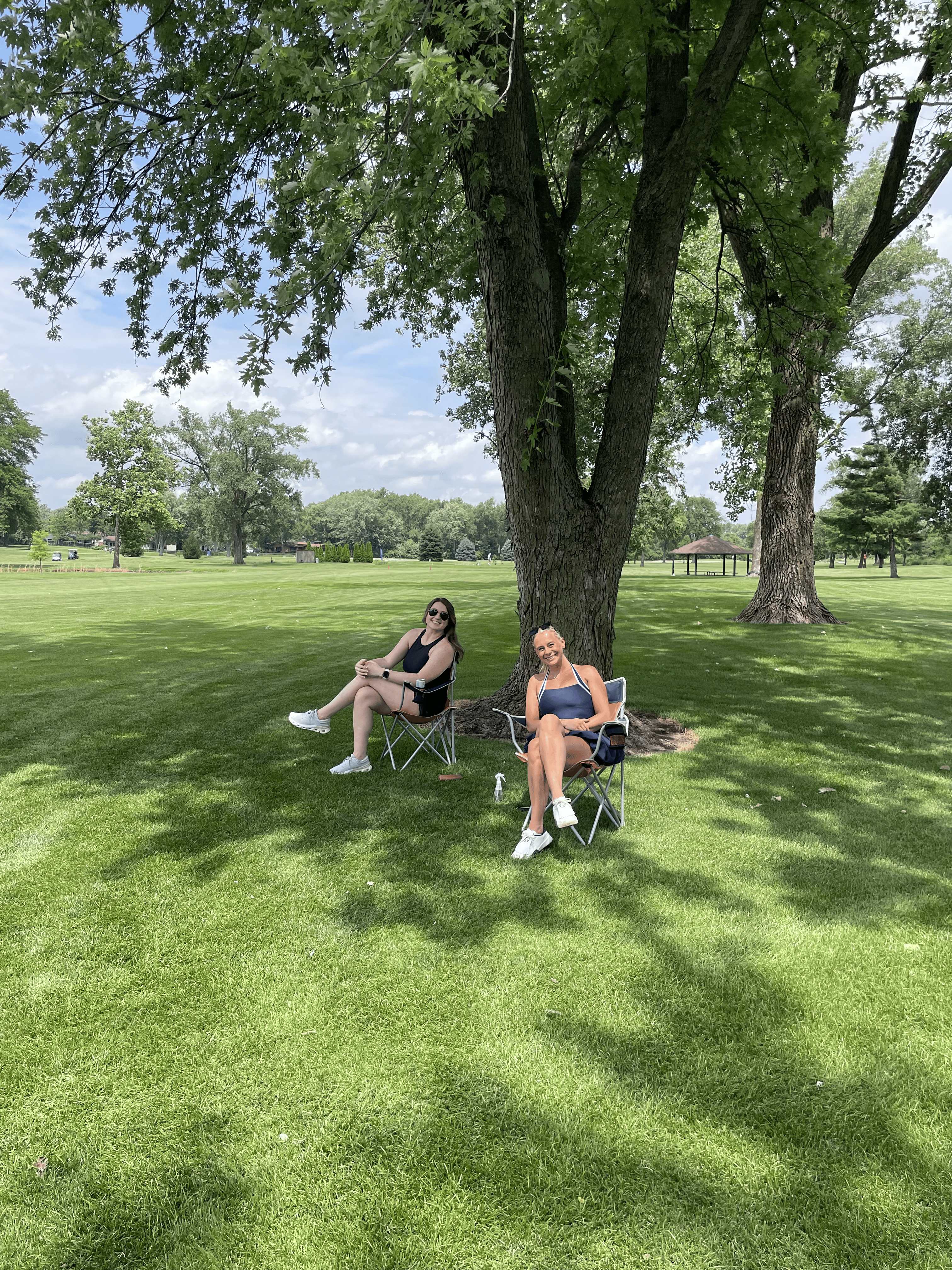 Two women relax in lawn chairs under a shade tree on the course during the inaugural outing